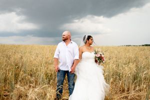 Newlyweds walking hand in hand through golden wheat field under stormy sky.