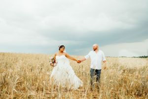 A couple holds hands in a golden wheat field on their wedding day against a cloudy sky.