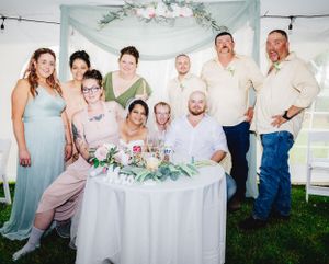 A wedding reception group gathers around a decorated table under a floral archway.