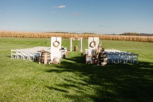 Outdoor wedding ceremony setup with white chairs and decorative pillars on a grassy field bordered by cornfields.