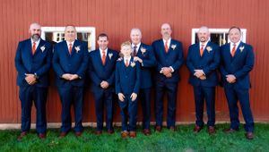 Groomsmen in navy suits with red ties lined up against red barn backdrop.
