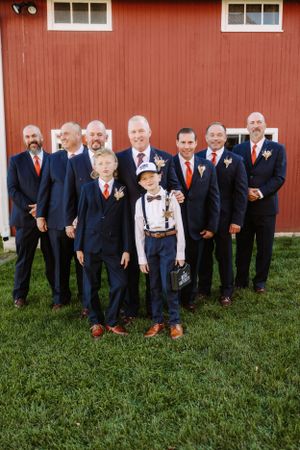 Wedding party in navy suits with orange accents posed in front of a red barn.