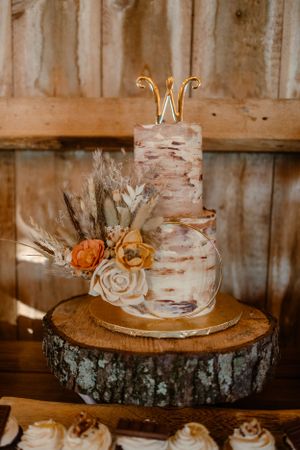 Two-tier birch bark wedding cake with dried flowers and monogram topper on wooden slice stand.