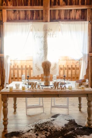 Elegant wedding sweetheart table with white curtains and decorative text display in a rustic barn.