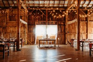 Light-filled barn interior set for wedding reception with wooden tables and hanging string lights.