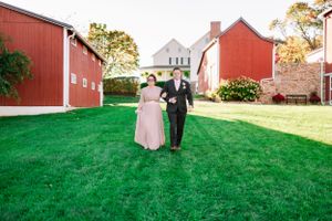 A couple walking together on green grass with red barns in the background.