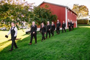Wedding party members walking in a line along a red barn on a sunny autumn day.