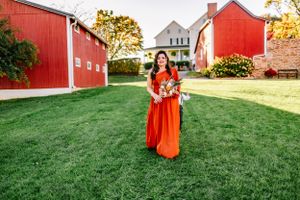 Woman in a vibrant red dress holding flowers poses in front of rustic red barns.