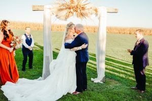 A wedding ceremony takes place under a rustic arch with cornfields in the background during golden hour.