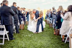 Wedding guests line up outdoors on white chairs while petals scatter across a grassy aisle at sunset.