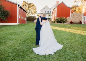 A romantic wedding portrait in front of red barns and a white farmhouse at sunset.