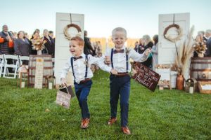 Two young ring bearers in suspenders and bow ties running across a grassy ceremony space.