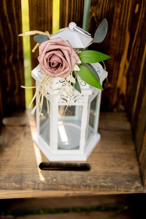 White decorative lantern adorned with a dusty pink rose and greenery on wooden surface.