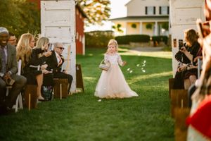 Flower girl in white dress walking down outdoor aisle with petals.
