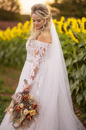 Bride in lace gown poses with fall-themed bouquet against backdrop of sunflowers at sunset.