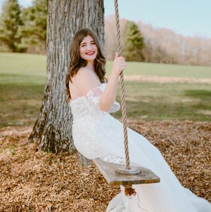 A joyful bride in a white dress swings on a rope swing near a large tree.
