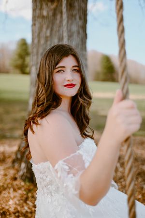 A bride in lace dress poses outdoors near a wooden swing on an autumn day.