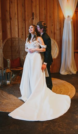 Wedding couple sharing a joyful moment in rustic barn venue with peacock chair and white drapes.