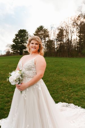 A bride in a lace detailed ball gown poses outdoors holding a white floral bouquet.