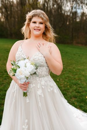 A radiant bride in a flowing lace gown holds a white bouquet against a green outdoor backdrop.