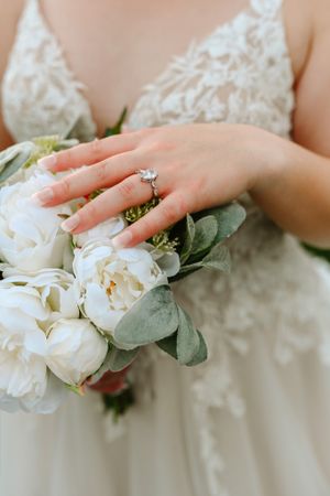 Close-up of a bride's hand with engagement ring holding white peonies and eucalyptus.