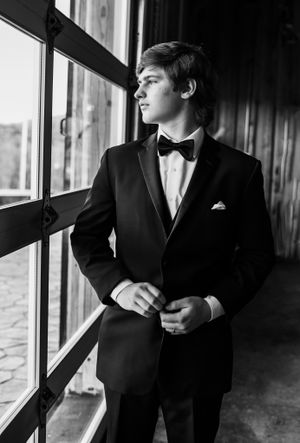A groom in classic black tuxedo gazes contemplatively out a window in black and white.