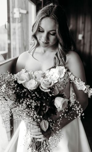 A bride in white dress admires her bouquet of roses and baby's breath in black and white.