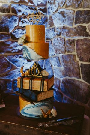 A Harry Potter themed wedding cake with golden details and Deathly Hallows symbols against stone wall.