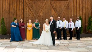 Wedding party in formal attire poses for photo against rustic barn backdrop with stone patio.