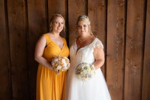 A bridesmaid in yellow and bride in white pose with bouquets against wooden barn wall.