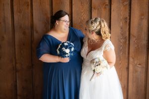 Bridesmaid in navy blue and bride in white gown share a candid moment against wooden wall.