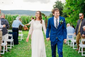 Newlyweds exit their outdoor ceremony on a grassy hill with white chairs and cornfields in background.
