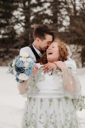 Joyful winter wedding portrait with blue bouquet and white dress with green accents.