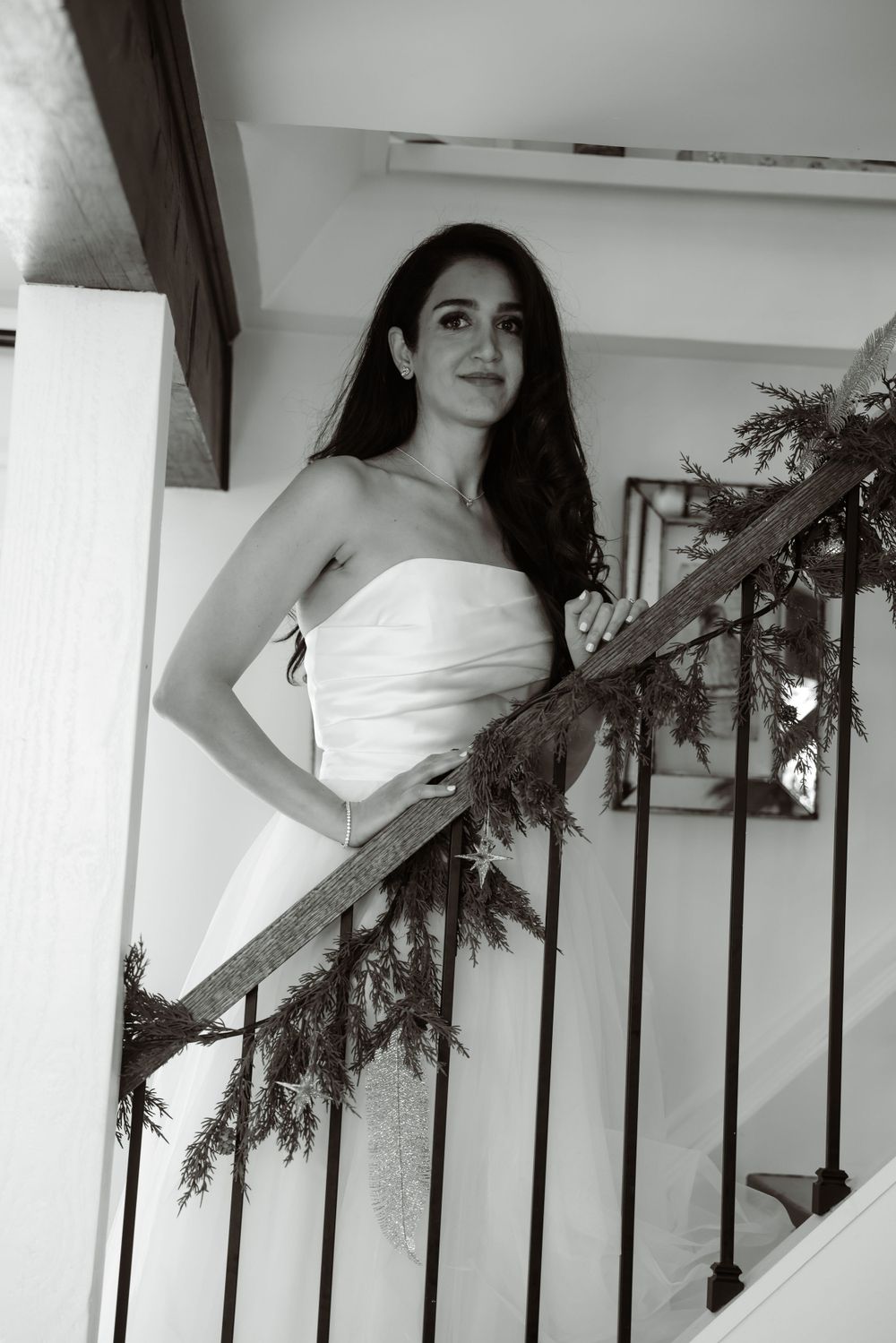 Black and white photo of a bride in strapless gown posing by a decorated staircase railing.