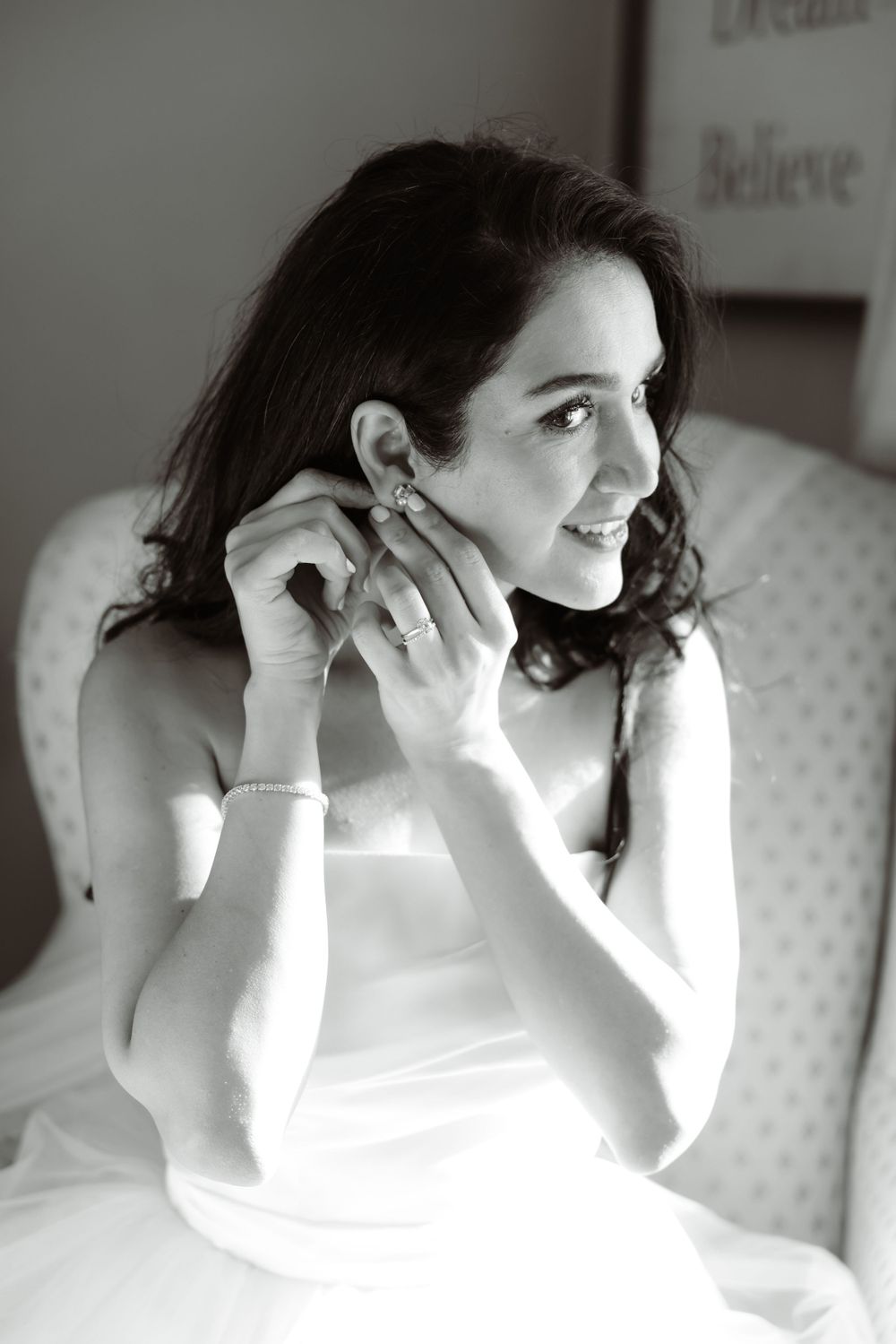 Black and white portrait of bride putting on earrings while seated in armchair.