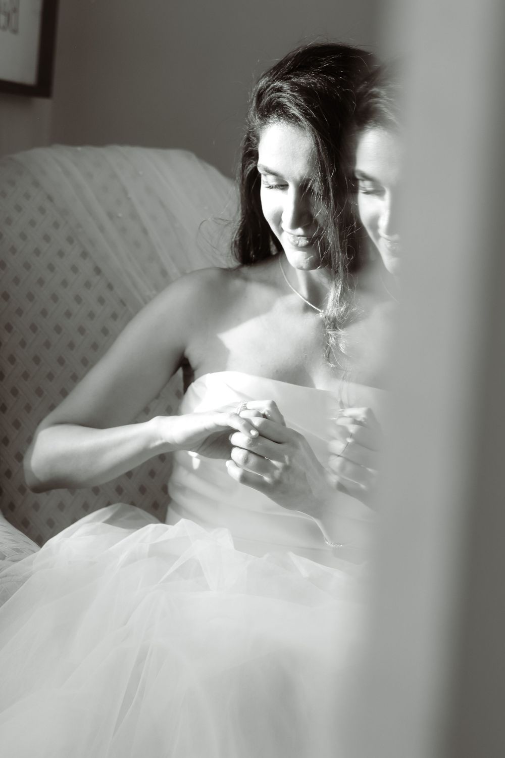 Artistic black and white photo through glass showing someone in bridal attire seated in chair.