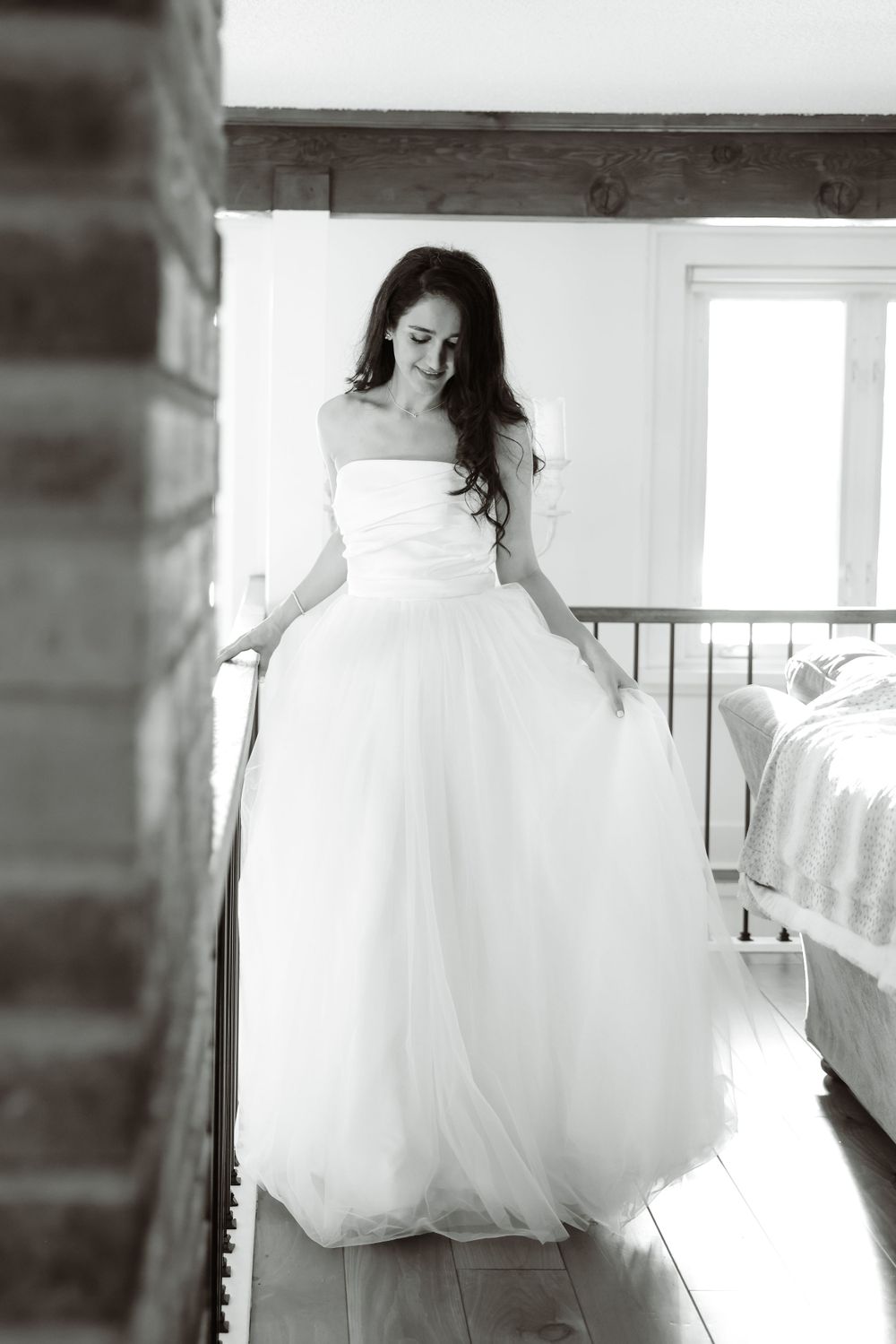 Black and white portrait of bride in flowing white gown standing by railing in natural light.