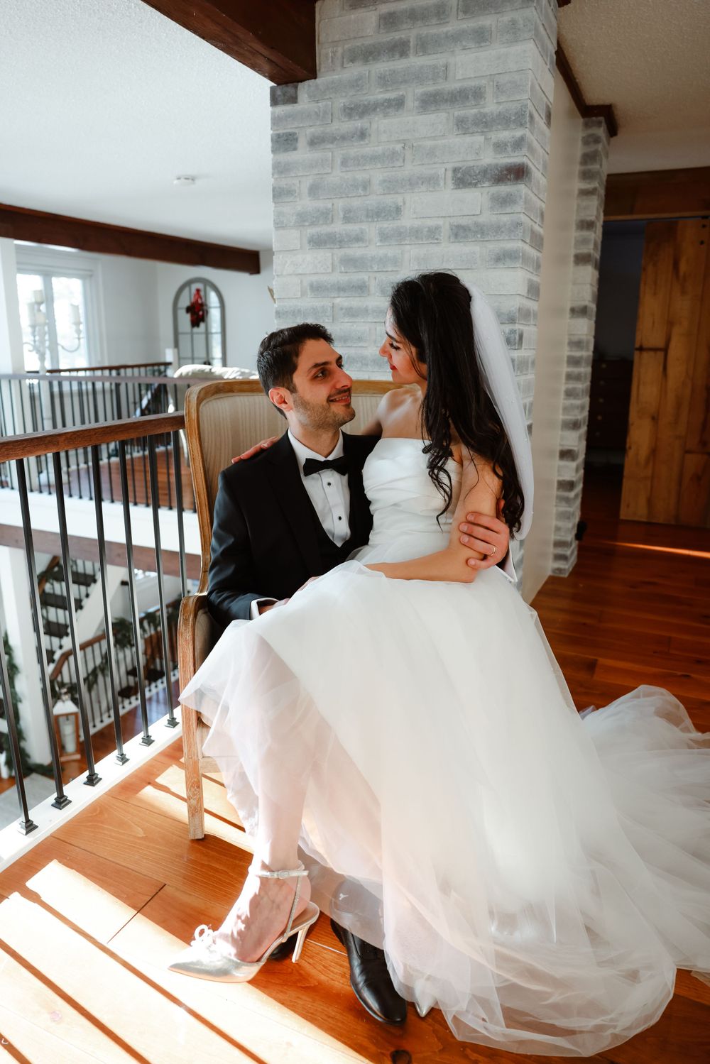 Sunlit wedding portrait on staircase landing with gray brick wall and wooden beams.