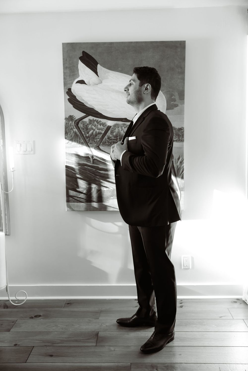 A groom in a classic black tuxedo adjusts his attire while standing in front of an abstract artwork.