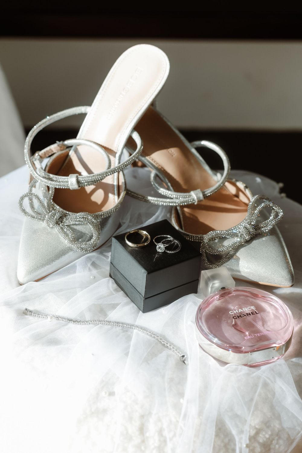 Elegant bridal shoes and wedding rings displayed with pink Chanel perfume on white tulle.