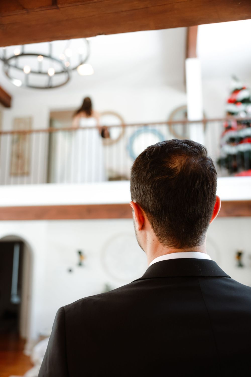 Back view of groom in black suit standing in elegant white interior with holiday decorations.