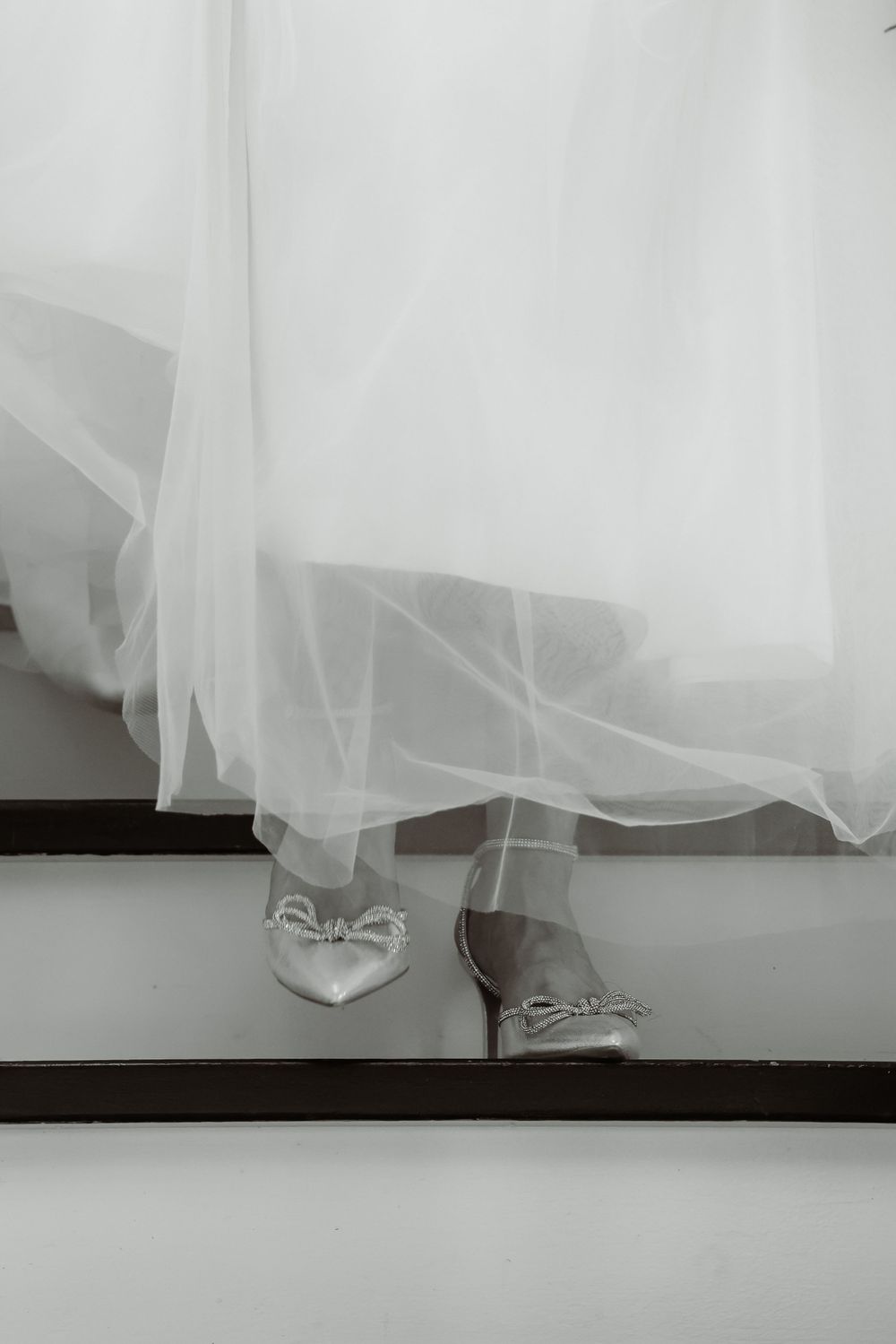 White bridal shoes peek out under flowing tulle wedding dress on wooden stairs.