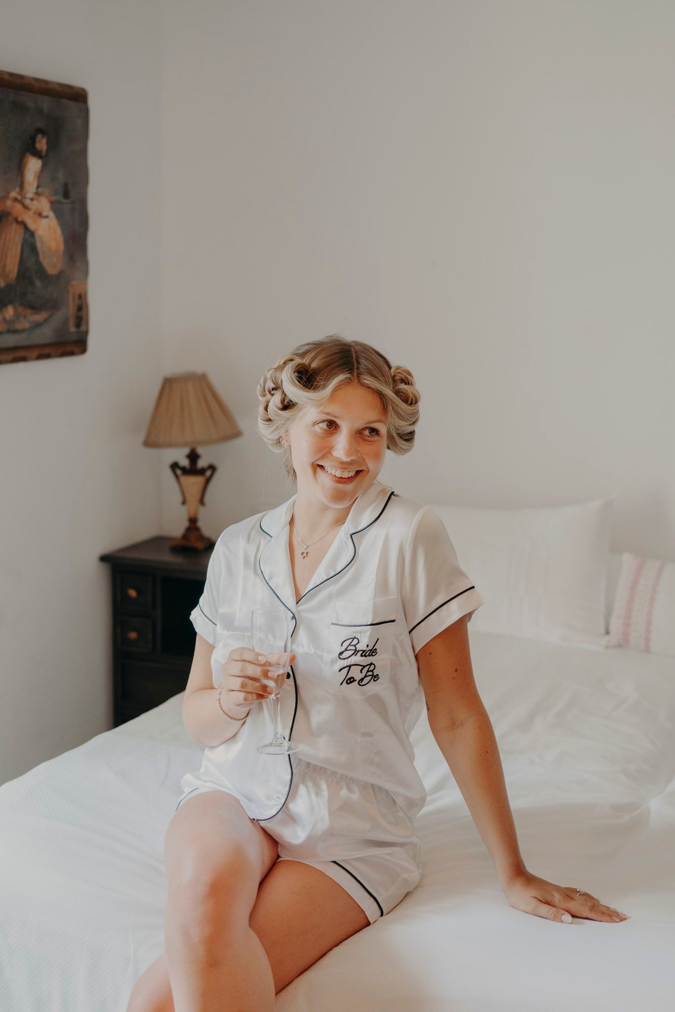 Vintage-style bedroom scene with person in white monogrammed pajama set with hair in curlers sitting on white bed.