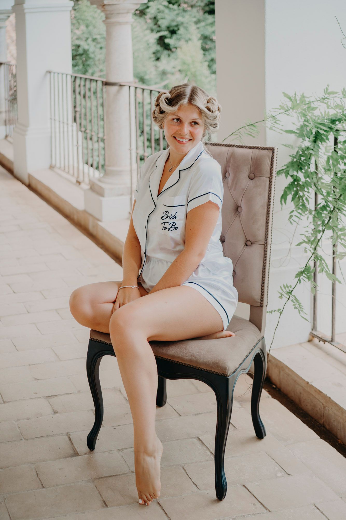 Person in white piped pajamas and hair curlers poses on tufted chair on outdoor porch with columns.