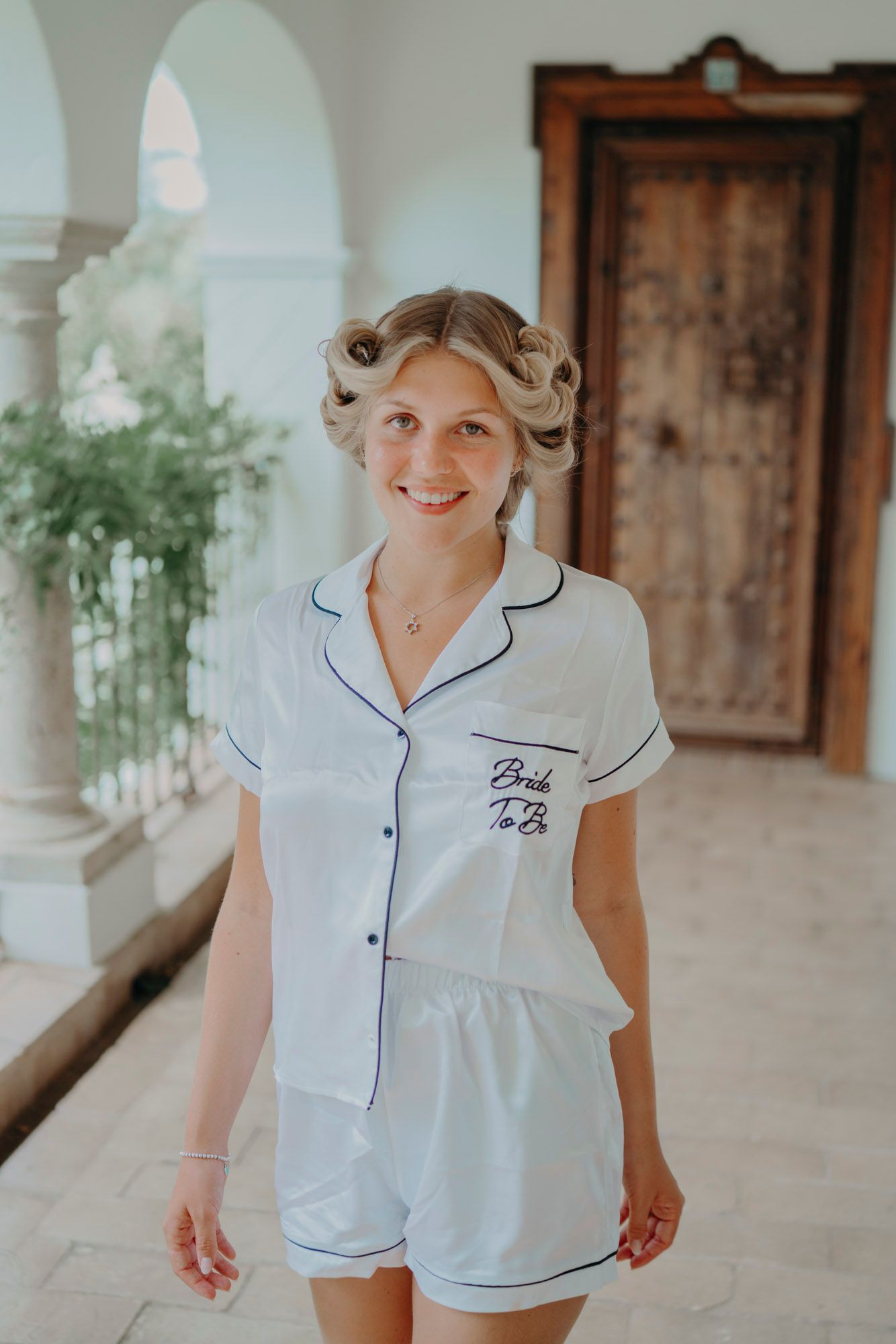 White bridal pajama set with navy piping trim and "Bride to Be" embroidery shown in elegant corridor setting.