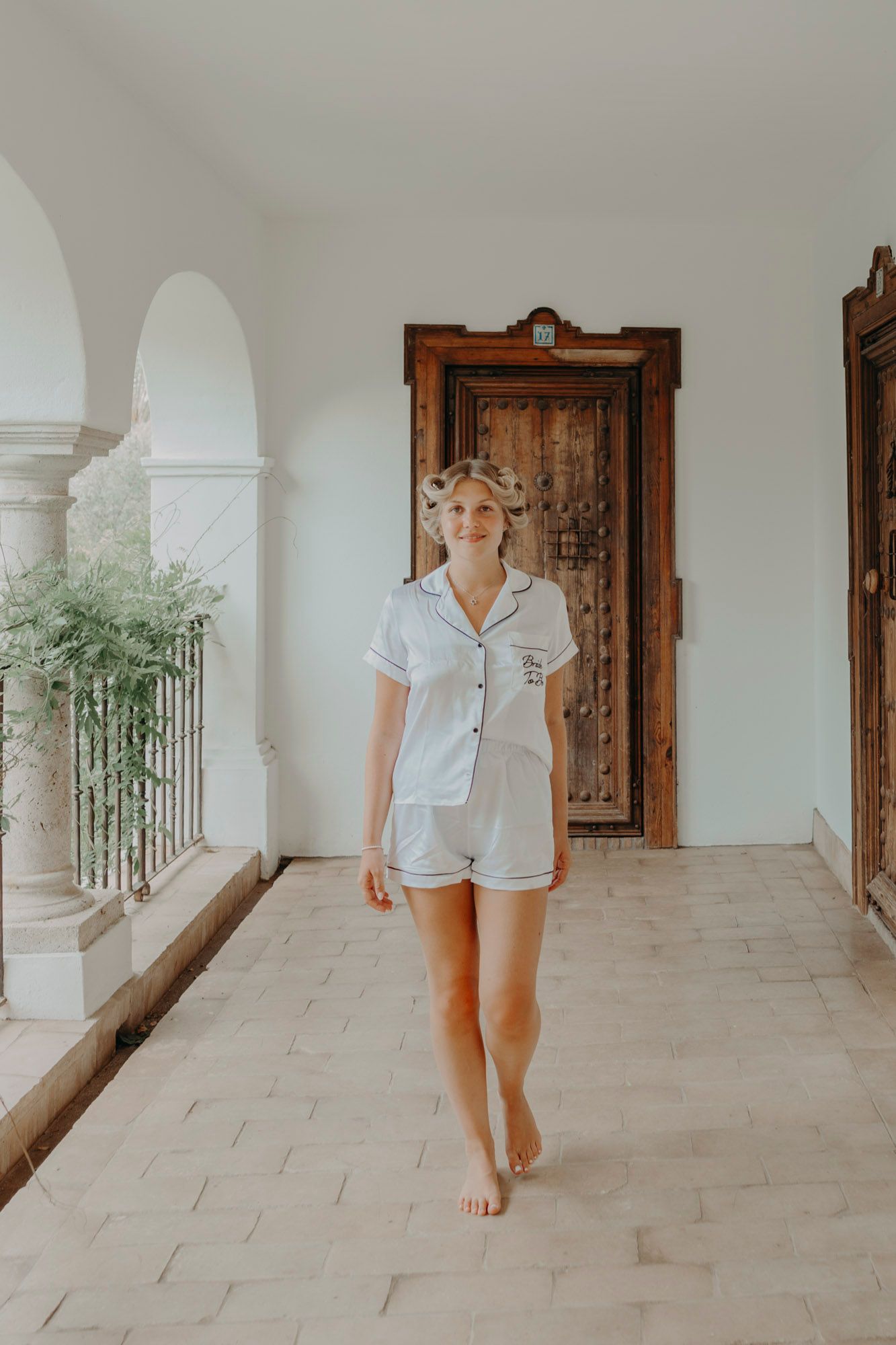 Someone wearing a crisp white cotton loungewear set walks barefoot along an elegant columned veranda with vintage wooden doors.