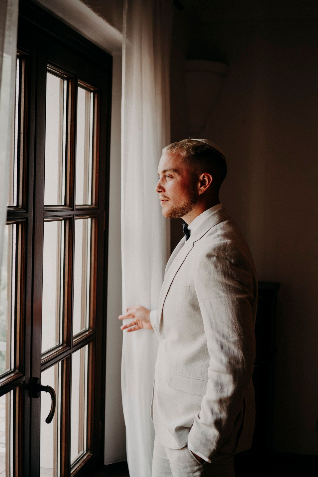 A person in a light gray suit stands contemplatively by a window with sheer curtains in moody lighting.