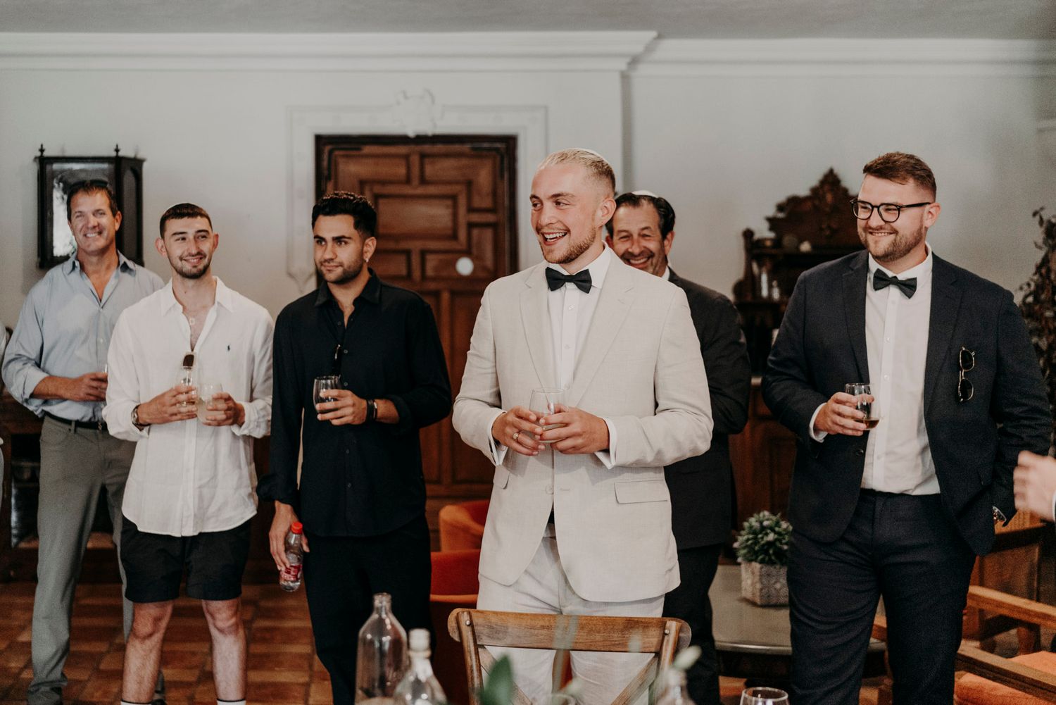 A group of well-dressed men in formal attire gather together holding drinks at an elegant indoor event.
