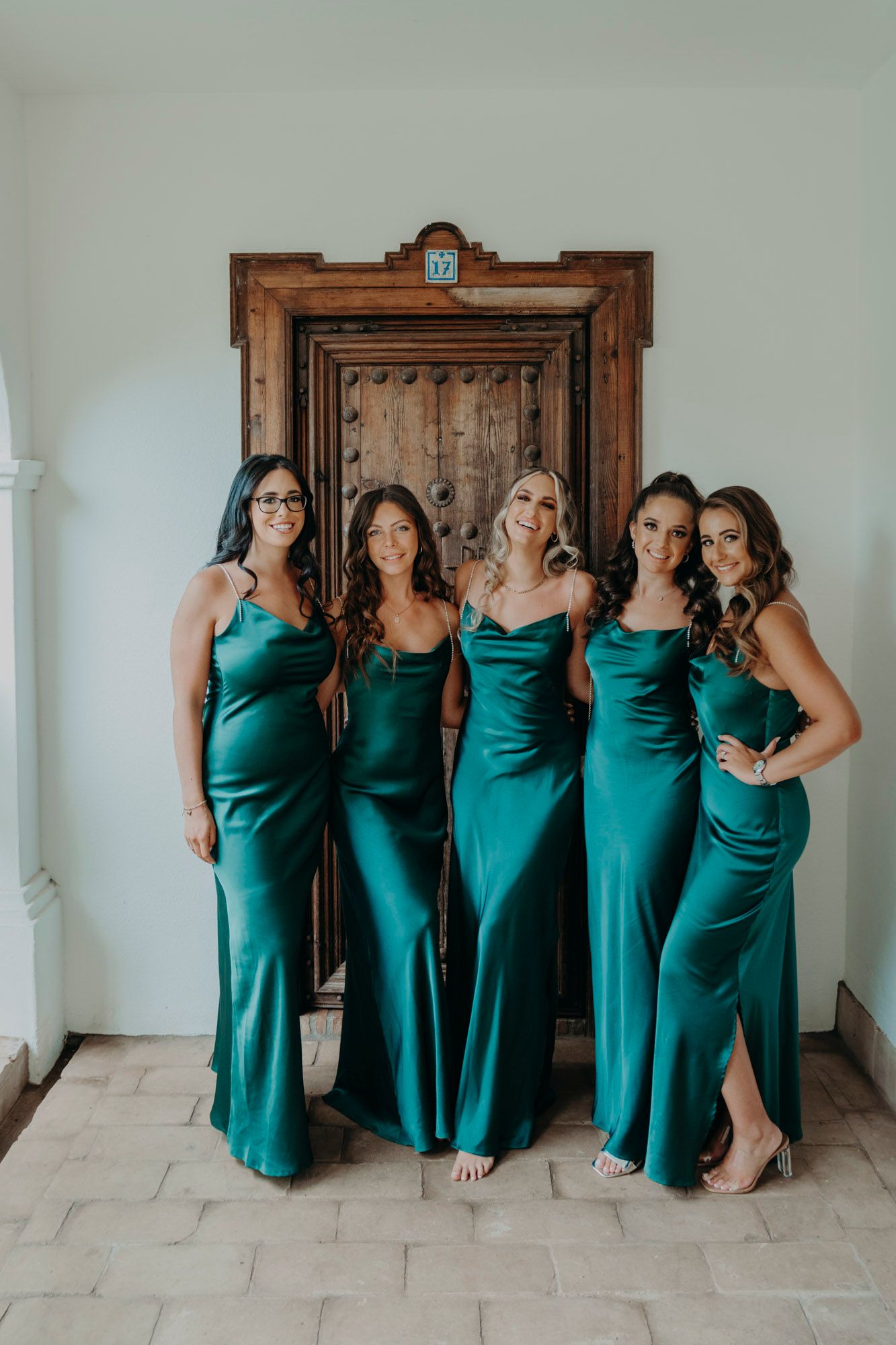 Five bridesmaids pose together in matching emerald green satin dresses in front of a rustic wooden door.