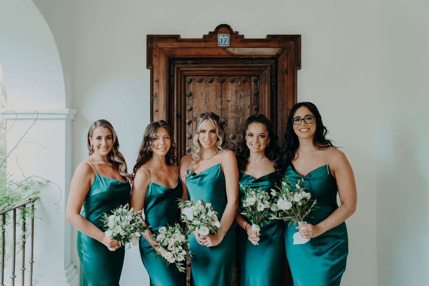 Bridesmaids in matching teal satin dresses pose together holding white flower bouquets on a bright porch.